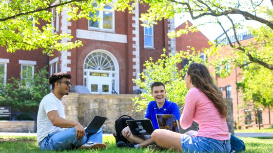 Three Martin School students sitting in the grass, laughing and talking, outside of Miller Hall.